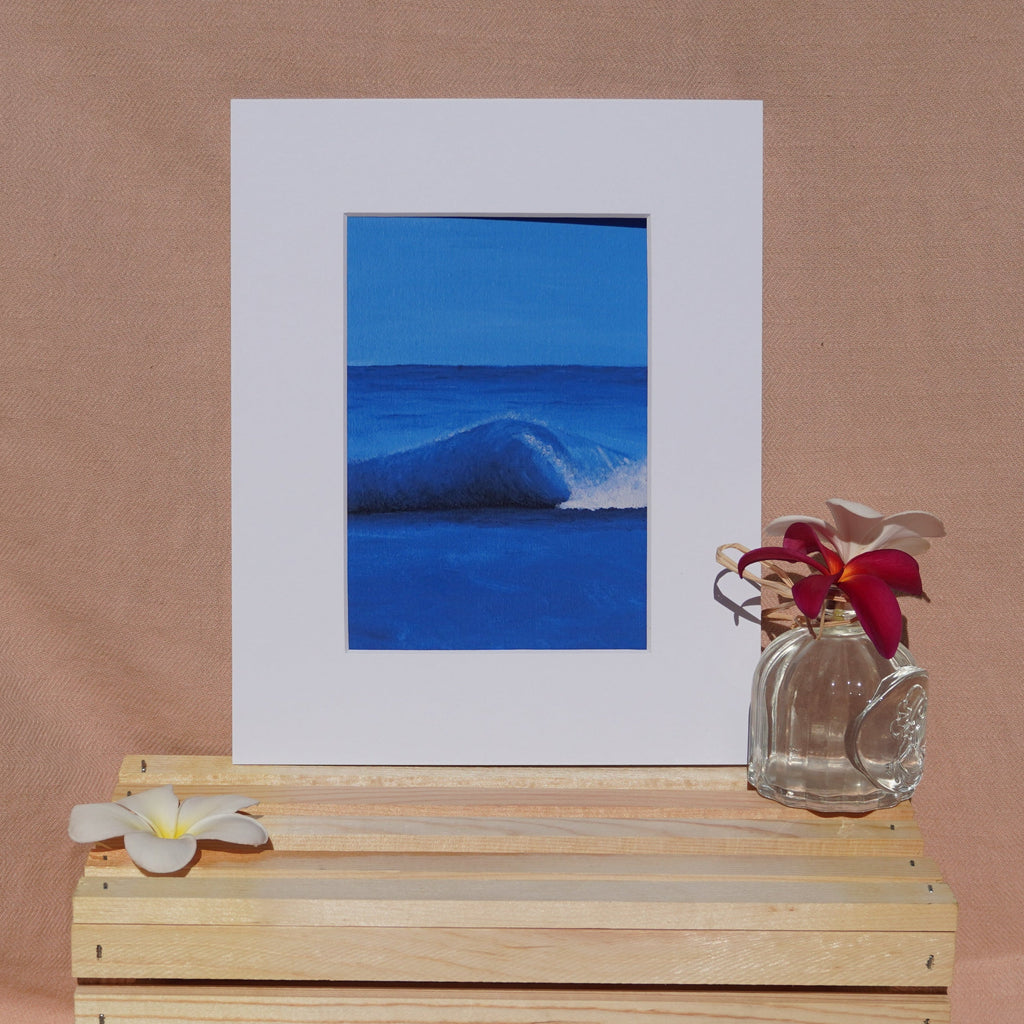A framed art print featuring a vibrant blue ocean wave, styled on a light wood crate with white plumeria flowers against a warm beige fabric backdrop.