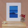 A framed art print featuring a vibrant blue ocean wave, styled on a light wood crate with white plumeria flowers against a warm beige fabric backdrop.