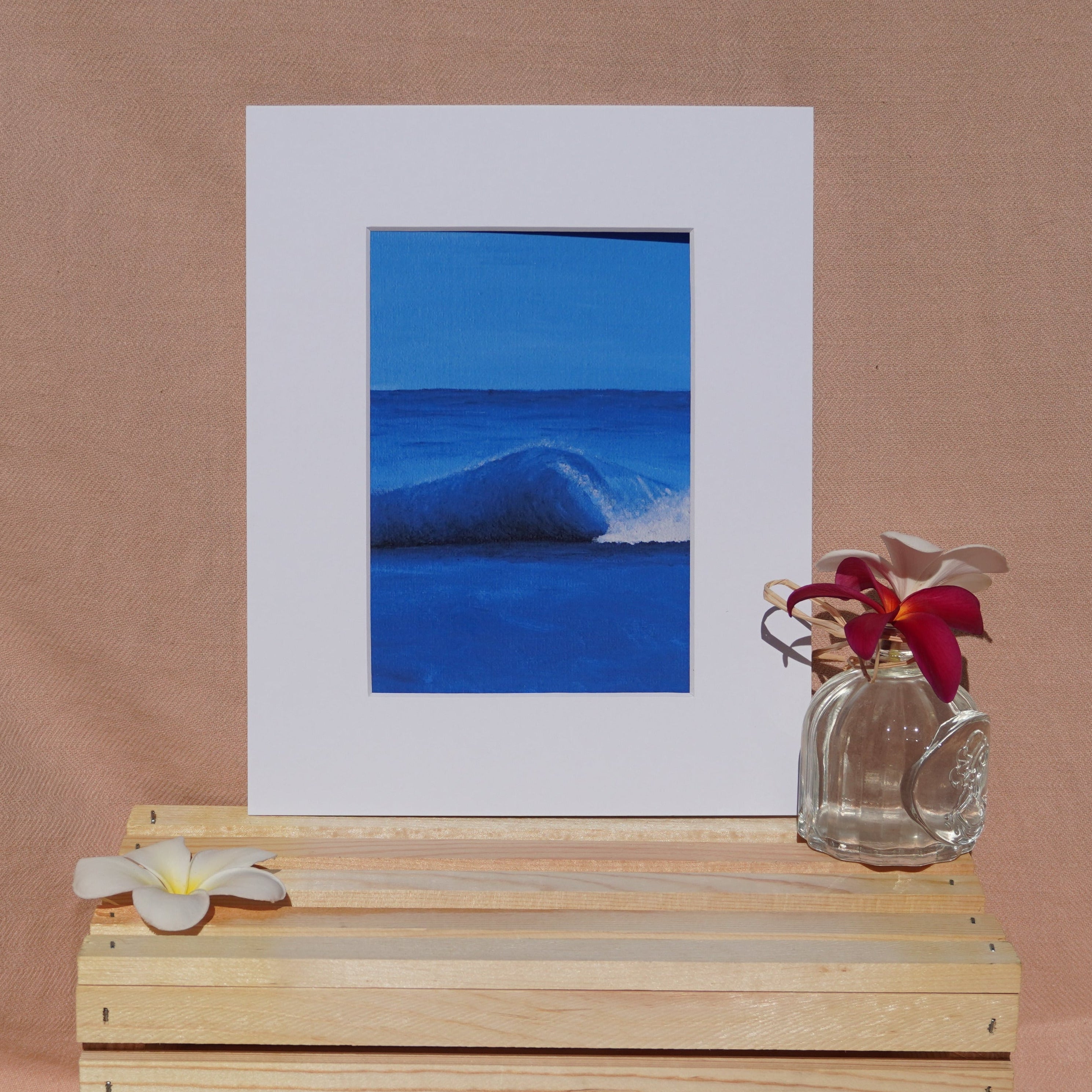A framed art print featuring a vibrant blue ocean wave, styled on a light wood crate with white plumeria flowers against a warm beige fabric backdrop.